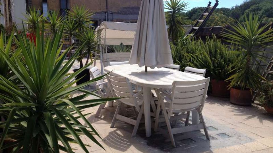 Terrazza pavimentata all’aperto con tavolo ovale e sedie bianche in plastica, ombrellone chiuso al centro e lettini sullo sfondo. Numerose piante in vaso con foglie appuntite circondano l’area, con edifici e tetti visibili oltre la ringhiera.