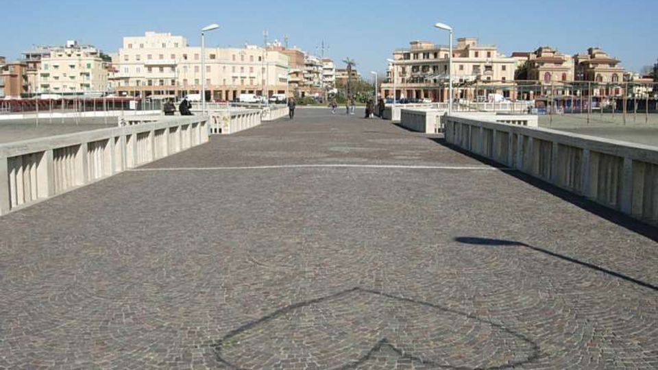 Ampio ponte pedonale lastricato in pietra, con parapetti in cemento e lampioni lungo i lati, che conduce verso un’area urbana costiera. Sullo sfondo si vedono edifici residenziali e commerciali di media altezza sotto un cielo sereno.