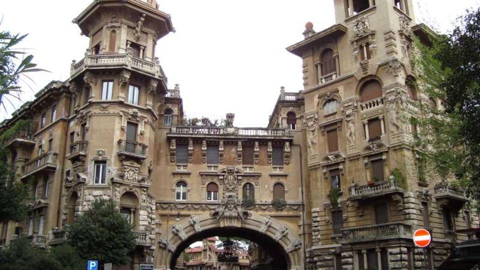 Palazzo storico in stile eclettico con facciate riccamente decorate, due torri angolari e un grande arco centrale che attraversa la strada. In primo piano si vedono alberi, auto parcheggiate e segnaletica stradale in un contesto urbano.