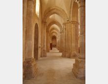 Interno di un chiostro o corridoio in pietra con lunga fila di colonne massicce e archi a tutto sesto, che crea una prospettiva profonda. Pavimento in lastre chiare e volta in muratura, con nicchie e aperture laterali lungo le pareti.