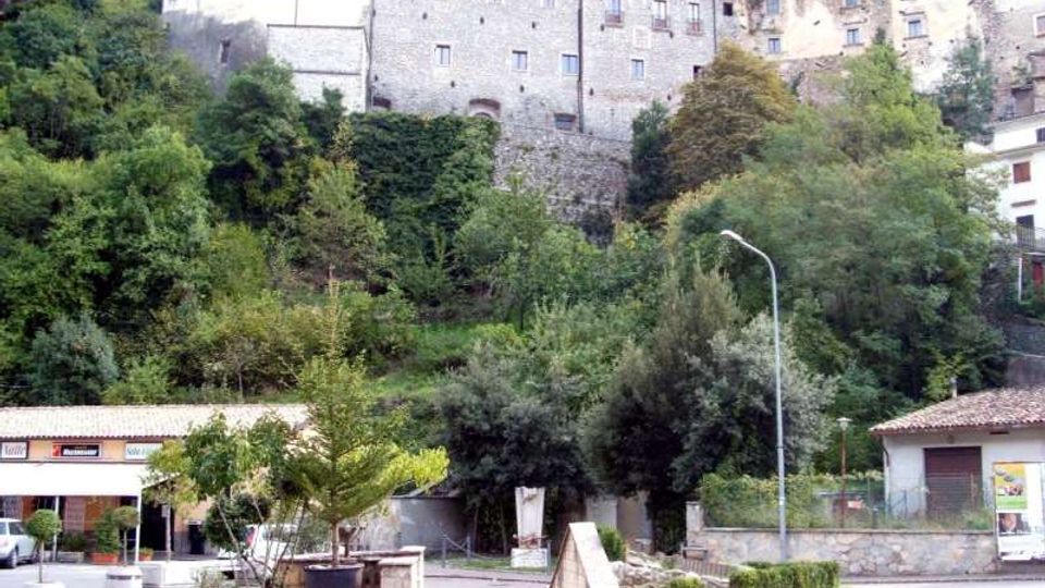 Veduta di un borgo collinare con case in pietra e intonaco addossate al pendio, sopra una fitta vegetazione di alberi e cespugli. In primo piano si vede una piccola piazza con aiuole, una panchina, muretti in pietra e un lampione.
