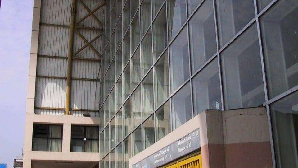 Facciata di un grande edificio moderno con parete vetrata a griglia e ampia pensilina metallica sostenuta da travi. Al piano strada si vedono ingressi con cancelli gialli e un porticato lungo il marciapiede in ambiente urbano.