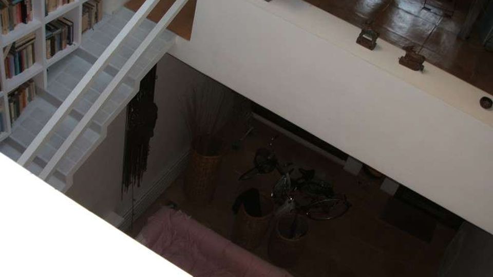 Interno di una casa su due livelli con soppalco e scala bianca; a sinistra c’è una grande libreria a parete e sotto si intravede una zona giorno con divano e vasi decorativi. Pavimento in legno e parapetti bianchi definiscono lo spazio aperto.