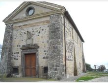 Facciata di una piccola chiesa in pietra e intonaco, con frontone triangolare e finestra circolare in alto, portale centrale in pietra e porta in legno. L’edificio è isolato su un’area erbosa, con muri laterali in muratura a vista e pochi elementi decorativi.