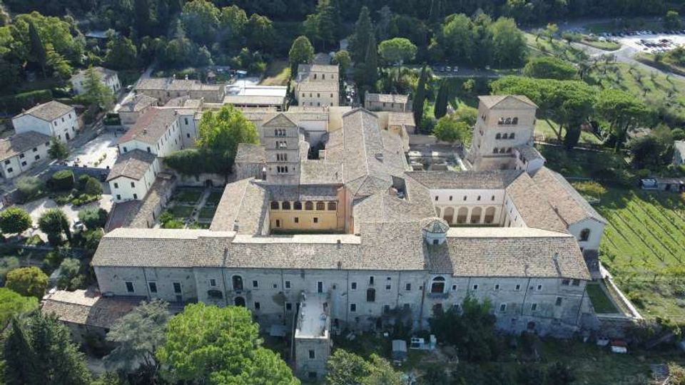 Veduta aerea di un grande complesso monastico in pietra con tetti in coppi, disposto attorno a cortili interni e con una torre campanaria. L’edificio è circondato da alberi e aree verdi, con campi coltivati e parcheggi nelle vicinanze.