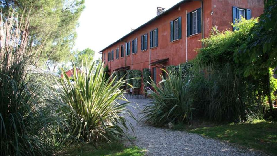 Edificio rurale a due piani con facciata intonacata color terracotta e persiane scure, affacciato su un vialetto in ghiaia. Intorno si trova un giardino con cespugli fitti, piante ornamentali e alberi alti che incorniciano l’ingresso.
