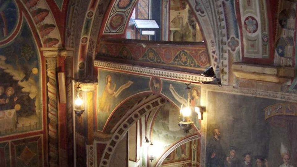 Interno di una chiesa o cappella con archi sovrapposti e soffitti a volta interamente decorati con affreschi e motivi geometrici. Una scala in pietra scende verso un livello inferiore illuminato da lampade a parete, con pavimento a mosaico e pannelli informativi.