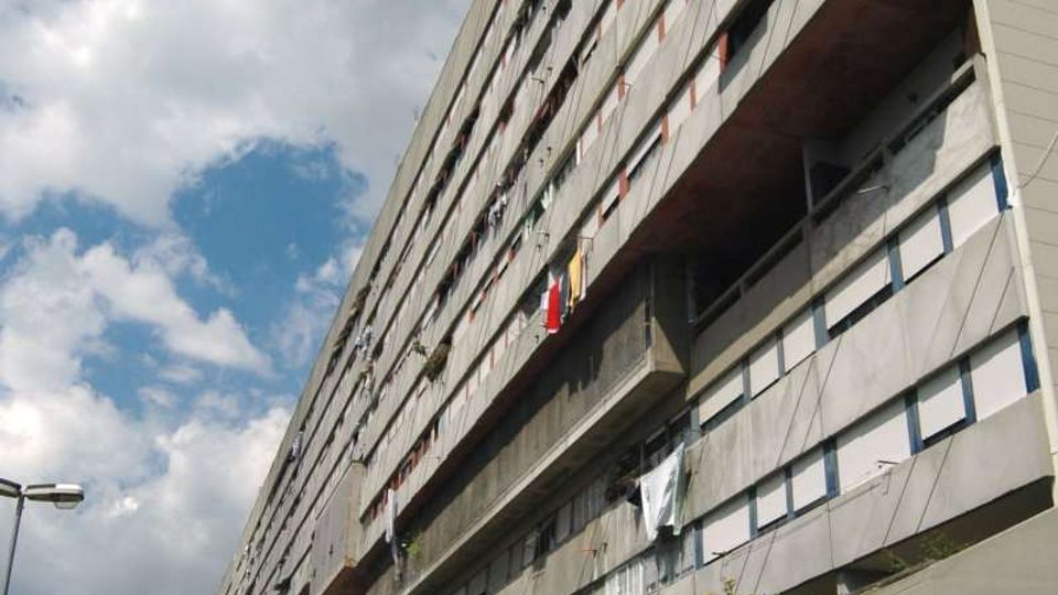 Grande edificio residenziale multipiano in cemento con lunga facciata lineare, file di finestre e ampi ballatoi incassati. In primo piano si vede una strada con auto parcheggiate, una rampa e scale in cemento, sotto un cielo parzialmente nuvoloso.