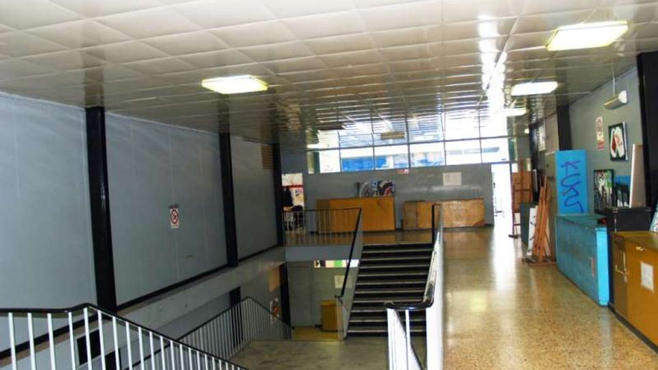 Interno di un edificio pubblico con ampio corridoio e pianerottolo che affaccia su una scala centrale, illuminato da plafoniere a soffitto. Pavimento in graniglia, pareti grigie con ringhiere metalliche e pannelli o bacheche lungo il lato destro.