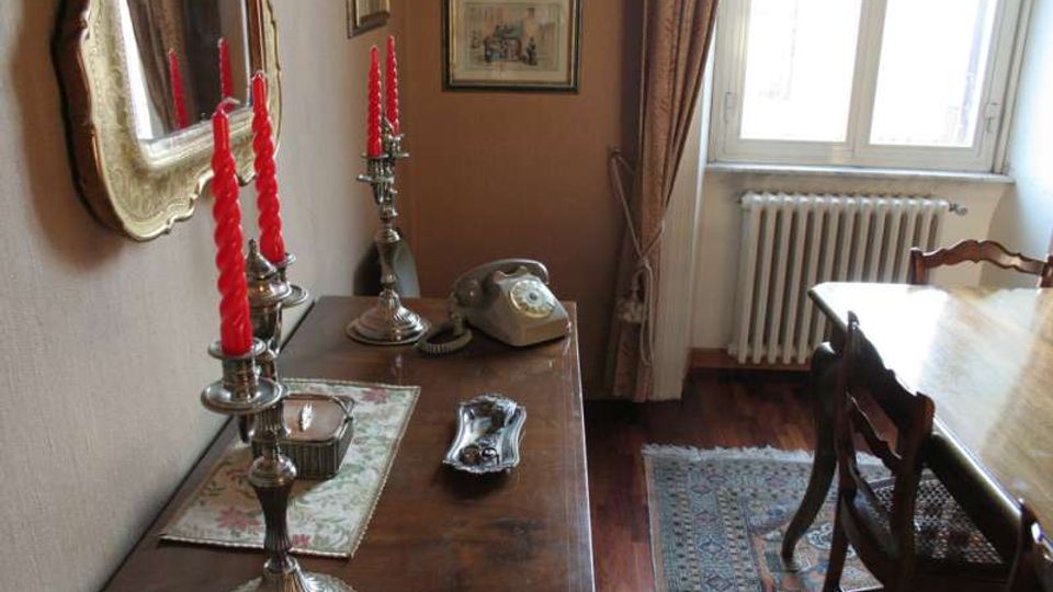 Interno di una sala da pranzo con tavolo in legno e sedie, finestra con tende e termosifone sotto. In primo piano una credenza con candelabri in argento e candele rosse, telefono d’epoca, specchio e quadri alle pareti, pavimento in legno con tappeto.