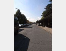 Strada urbana asfaltata e dritta con auto parcheggiate lungo entrambi i lati. A destra si vede una lunga tettoia con struttura metallica e alberi alti sullo sfondo, sotto un cielo sereno.