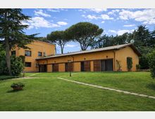 Edificio basso in intonaco giallo con tetto a falde e grandi porte in legno, affacciato su un ampio prato con vialetto in pietra e lampione. Sullo sfondo si vedono alti alberi mediterranei e un cielo azzurro con nuvole sparse.
