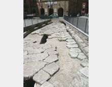 Antica strada lastricata in grandi pietre irregolari, incassata in una trincea e delimitata da muretti in pietra e ringhiere metalliche. Sullo sfondo si vede una piazza con auto parcheggiate e un edificio storico con archi e targhe sulla facciata.