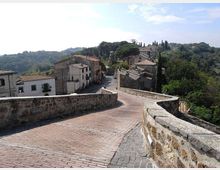 Strada in mattoni su un ponte o camminamento in pietra con muretti laterali, che scende verso un piccolo borgo con case in muratura e tetti in tegole. Sullo sfondo si vedono colline alberate con cipressi e vegetazione mediterranea sotto un cielo sereno.
