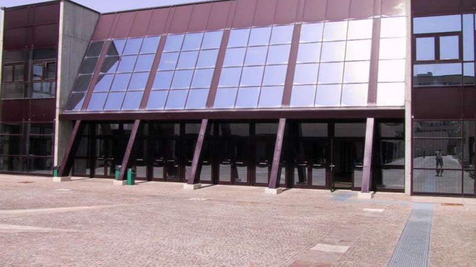 Facciata di un edificio moderno con ampia superficie vetrata inclinata e pannelli metallici, sostenuta da pilastri obliqui sopra un ingresso con porte scure. Davanti si apre una grande piazza pavimentata in pietra, con cielo sereno sullo sfondo.
