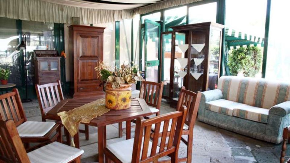 Veranda chiusa con grandi vetrate e tenda a soffitto, arredata con tavolo e sedie in legno, divano e mobili vetrina. Pavimento in pietra e piante decorative, con vista su area esterna ombreggiata da pergolato.