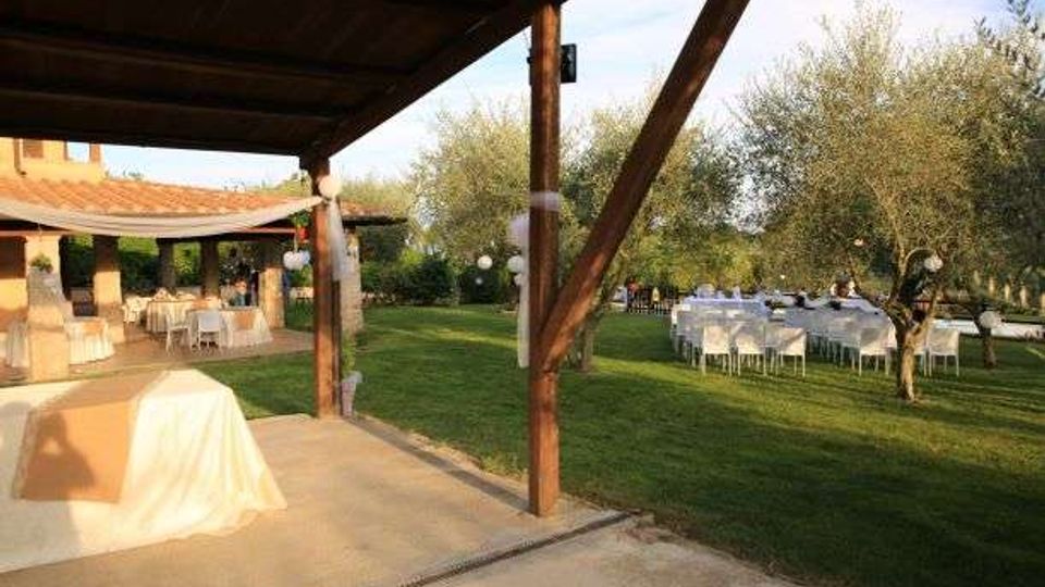 Spazio esterno per eventi con portico in legno e pavimentazione in cemento affacciato su un prato verde. Sul prato sono disposti tavoli e sedie bianche apparecchiati, circondati da ulivi e da un edificio con tetto in coppi e area coperta.