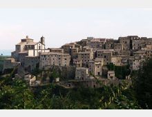 Borgo medievale su un pendio con fitte case in pietra dai tetti in coppi e una chiesa con campanile sul lato sinistro. In primo piano si vedono vegetazione e alberi, con colline sullo sfondo sotto un cielo chiaro.