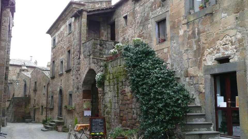 Stradina acciottolata in un borgo storico con edifici in pietra e intonaco, archi e balcone al piano superiore. Vasi con piante e un grande arbusto vicino a una scalinata esterna e a un ingresso con porta vetrata.