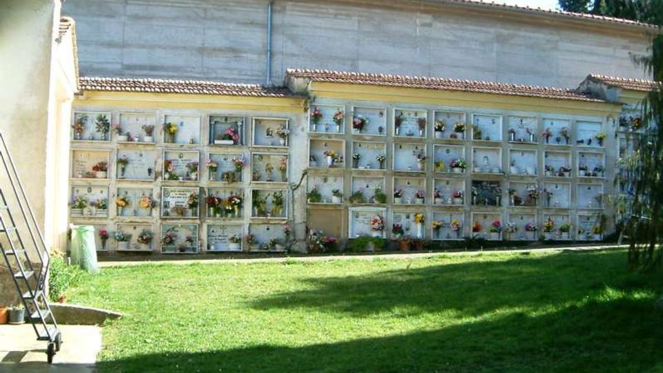 Cortile di un cimitero con prato verde e un lungo muro di loculi in cemento, disposti in file regolari e decorati con vasi di fiori. Sullo sfondo si vede l’edificio con tetto in tegole e alcuni alberi ai lati.