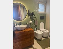 Bagno moderno con pareti verde salvia, mobile lavabo in legno con lavandino a ciotola e grande specchio rotondo retroilluminato. A destra si vedono bidet e WC sospesi sotto una finestra satinata, con accessori a parete e tappetino sul pavimento chiaro.