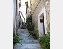 Vicolo stretto in un borgo con lunga scalinata in pietra che sale tra edifici addossati, muri in muratura e una porta incorniciata di bianco. Lungo i gradini ci sono vasi con piante verdi e lampioni a parete sospesi sopra il passaggio.