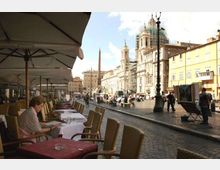 Piazza urbana con pavimentazione in ciottoli, tavolini e sedie di un caffè all’aperto sotto grandi ombrelloni lungo il bordo sinistro. Sullo sfondo si vede una chiesa barocca con cupola (parzialmente coperta da impalcature), un obelisco al centro della piazza e palazzi storici illuminati dal sole.