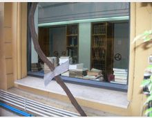 Finestra su strada che mostra l’interno di una biblioteca o archivio, con scaffali alti pieni di volumi e numerose pile di libri su un tavolo vicino al vetro. Davanti alla finestra passa una struttura metallica arrugginita e, sotto il davanzale, si vedono tubazioni esterne lungo la parete.