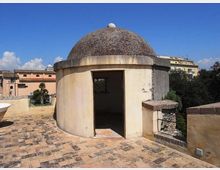 Piccolo edificio circolare in muratura con cupola scura e ingresso aperto, situato su una terrazza pavimentata in pietra. Sullo sfondo si vedono edifici cittadini, alberi e cielo sereno.