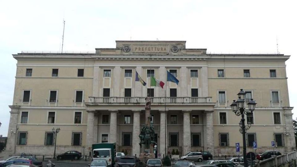 Edificio pubblico a più piani con facciata neoclassica, portico centrale con colonne e balcone, e frontone con la scritta “PREFETTURA”. Davanti si vede una piazza con auto parcheggiate, un monumento su piedistallo e lampioni stradali.