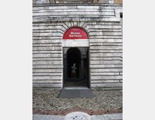 Ingresso del Museo Barracco con portale ad arco in pietra bugnata e insegna rossa sopra l’architrave. All’interno del corridoio si intravede una statua, con pavimentazione in lastre di pietra e mattoni davanti all’entrata.