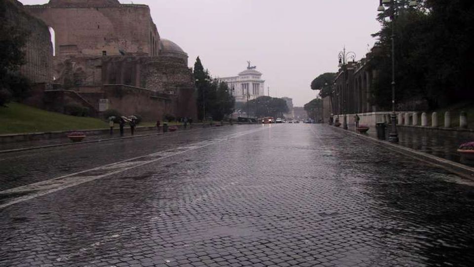 Ampio viale urbano in pavé bagnato dalla pioggia, fiancheggiato da alberi e lampioni, con i resti di un grande edificio antico in mattoni sulla sinistra. Sullo sfondo si vede un monumento bianco con statue in cima.