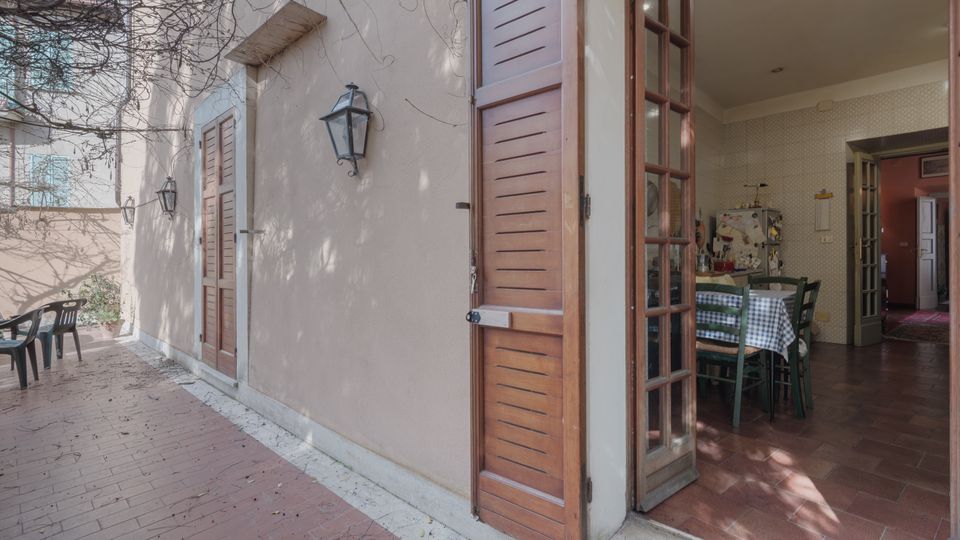 Cortile esterno pavimentato in mattoni lungo la facciata di una casa intonacata, con persiane in legno e lanterne a parete. Una portafinestra aperta collega al soggiorno con tavolo da pranzo e pavimento in cotto; sul fondo si intravede un corridoio interno.