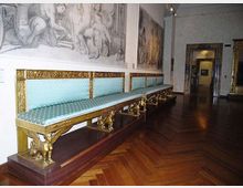 Sala museale con lungo divano imbottito azzurro e decorazioni dorate, sostenuto da piedini scolpiti, su pavimento in parquet a spina di pesce. Alle pareti sono visibili grandi disegni murali e, in fondo, un passaggio ad arco verso un’altra stanza.