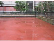 Campo sportivo all’aperto con pavimentazione rossa bagnata dalla pioggia e pozzanghere, circondato da recinzioni metalliche. Sullo sfondo si vedono alberi e un edificio con facciata a pannelli bianchi e rossi.