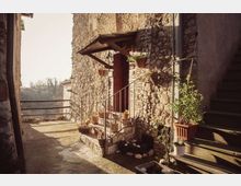 Cortile esterno in un borgo, con edificio in pietra e piccola veranda sopra l’ingresso raggiunto da pochi gradini con ringhiera in ferro. Vasi di piante lungo il muro e su una scala laterale, con parapetto sullo sfondo affacciato su una valle.