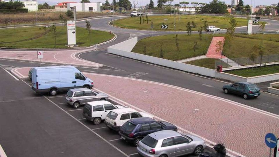 Rotatoria stradale moderna con aiuole erbose e giovani alberi al centro, circondata da carreggiate asfaltate e cartelli direzionali. In primo piano si vede un parcheggio con diverse auto e un furgone, con edifici bassi sullo sfondo.