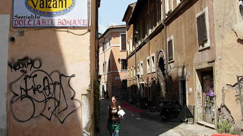 Vicolo stretto con pavimentazione in ciottoli tra edifici in muratura color ocra con persiane, finestre e segni di usura. In primo piano c’è un grande cartello “Pasticceria Valzani” su una parete con graffiti, mentre lungo la strada si vedono un portone ad arco e alcuni oggetti appoggiati ai muri.
