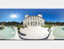 Veduta panoramica di un palazzo storico a pianta curva, con facciata chiara e finestre regolari, affacciato su un ampio viale in ghiaia. In primo piano ci sono due vasche d’acqua e, tutt’intorno, un giardino formale con siepi geometriche, statue e alberi sullo sfondo.
