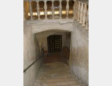 Scala interna in pietra che scende verso un ambiente sotterraneo con soffitto ad arco; in alto si vede una balaustra con colonnine. In fondo c’è una pesante porta a grata in legno, con pareti in muratura grezza e illuminazione soffusa.