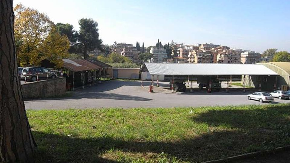 Area asfaltata con pensiline basse in metallo, simile a un parcheggio o deposito, vista da un prato in pendenza con alberi ai lati. Sullo sfondo si vede un quartiere collinare con edifici residenziali e vegetazione sotto un cielo sereno.