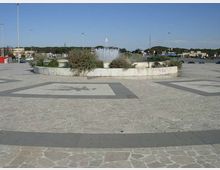 Ampia piazza pavimentata in pietra con motivi geometrici, al centro una fontana circolare rialzata con getti d’acqua e aiuole di vegetazione. Sullo sfondo si vedono lampioni alti, strade e costruzioni basse in un’area aperta.