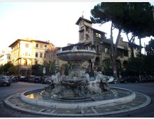 Fontana monumentale in una rotonda, con grande vasca circolare e sculture in pietra attorno al basamento. Sullo sfondo si vedono edifici storici con torretta e portici, alberi alti e auto parcheggiate lungo la strada.