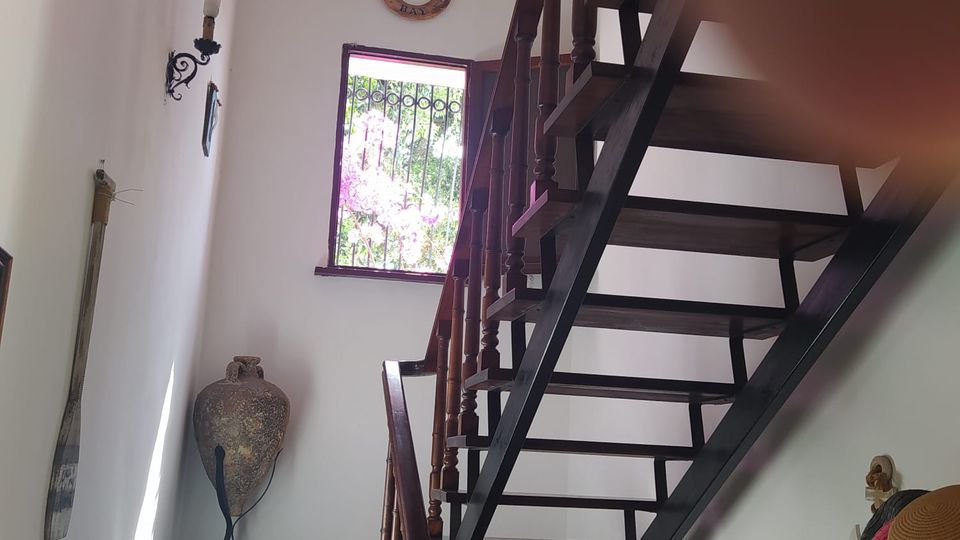 Interno di vano scala con pareti bianche e scala in legno scuro con struttura a gradini aperti e corrimano. Una finestra con grata in ferro lascia entrare luce naturale e sulla parete sono presenti una applique e un vaso decorativo su un piccolo pianerottolo.