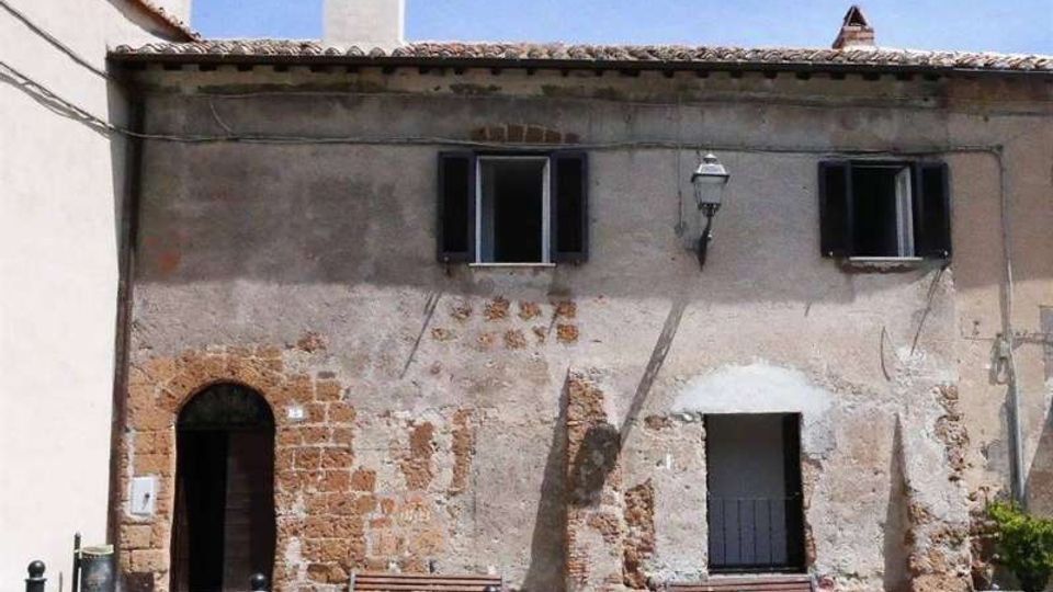 Facciata di un edificio in muratura intonacata e parzialmente scrostata, con tetto in coppi, comignolo e finestre con scuri scuri. Davanti si vede una piccola piazza lastricata con panchine in legno, paletti metallici e un lampione a parete.