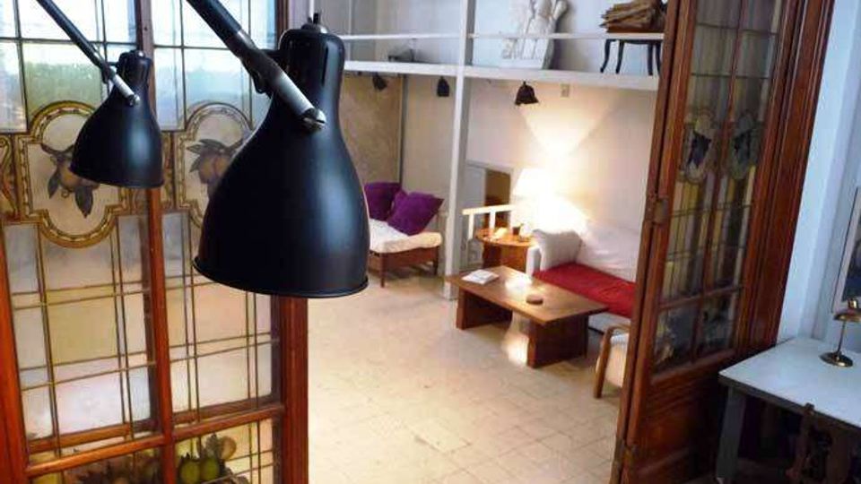 Interno di un loft con soffitti alti e soppalco con scaffalature bianche, affacciato su una zona giorno con divani e tavolino basso. In primo piano si vedono grandi lampade nere e una porta in legno con vetrate decorate che separa l’ambiente.