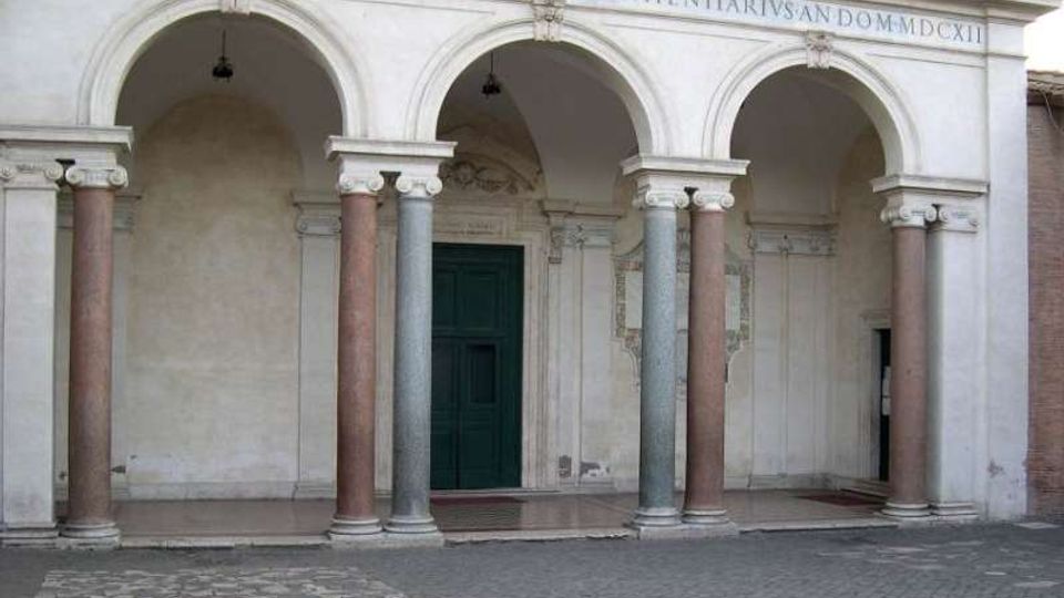 Portico in pietra con tre grandi archi a tutto sesto sostenuti da colonne in marmo di colori diversi, con iscrizione sulla trabeazione e lampade sospese. Al centro si vede una grande porta verde incorniciata da decorazioni, affacciata su un cortile pavimentato in pietra.