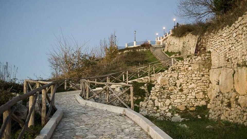 Sentiero in pietra in salita con parapetti in legno, che serpeggia tra muretti a secco e terrazzamenti erbosi su un pendio. In alto si intravedono lampioni e costruzioni lungo il percorso, con vegetazione rada ai lati.