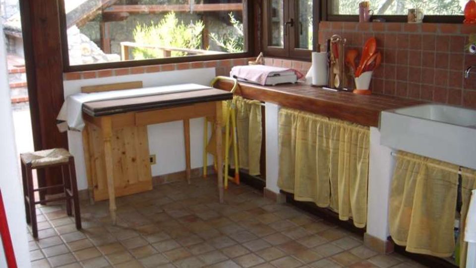 Cucina rustica con grandi finestre in legno che affacciano sul verde, pavimento in piastrelle e piano di lavoro in legno lungo la parete. Sotto il piano e il lavello ci sono tendine gialle, con paraschizzi in mattonelle e utensili da cucina appoggiati sul bancone.