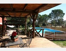 Veranda in legno coperta con tavoli e sedie da esterno, affacciata su una piscina rettangolare recintata con staccionata in legno. Sullo sfondo si vedono prato e alberi alti in un’area verde.